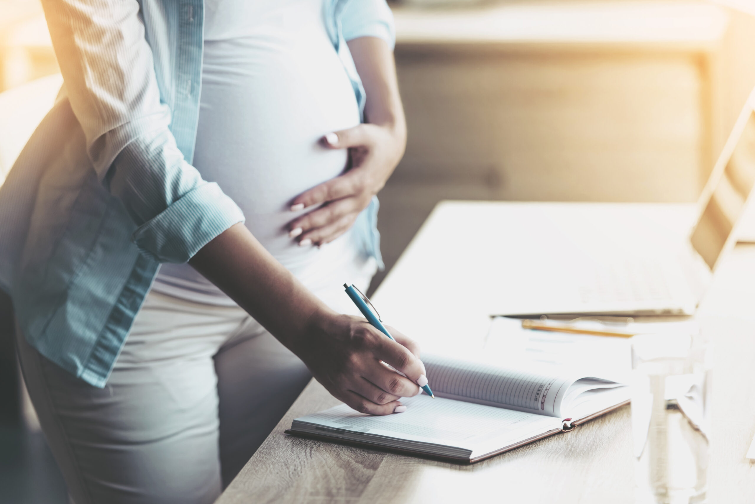 Pregnant Woman Writing in Notebook and Smiling while Standing near Working Place at Home. Freelancing at Home. Laptop on Desk. Remote Work Concepts. Wooden Table in Room.